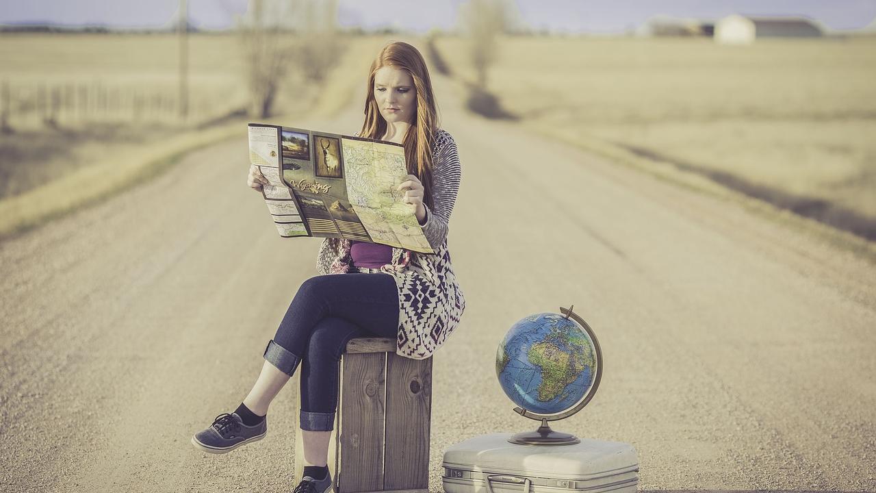 A white woman sits on a suitcase in the middle of the road contemplating a map while a world globe sits on top of a smaller suitcase next to her.