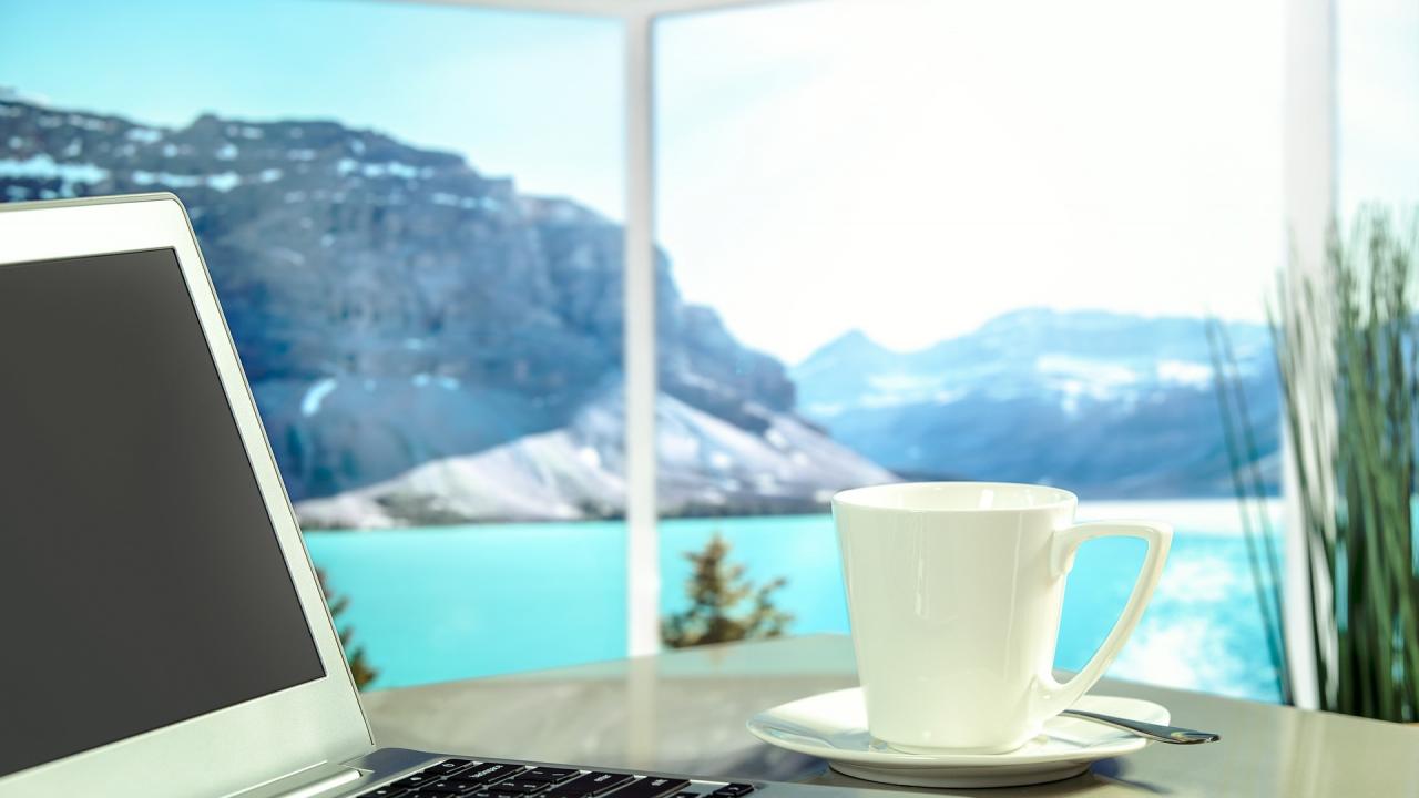 A laptop and coffee sit in the foreground. Beyond is a large window overlooking a lake with mountains in the background.