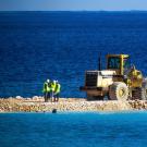 Three construction workers conferring next to a bulldozer, all of which stand on a small man-made gravel island, surrounded by the sea.