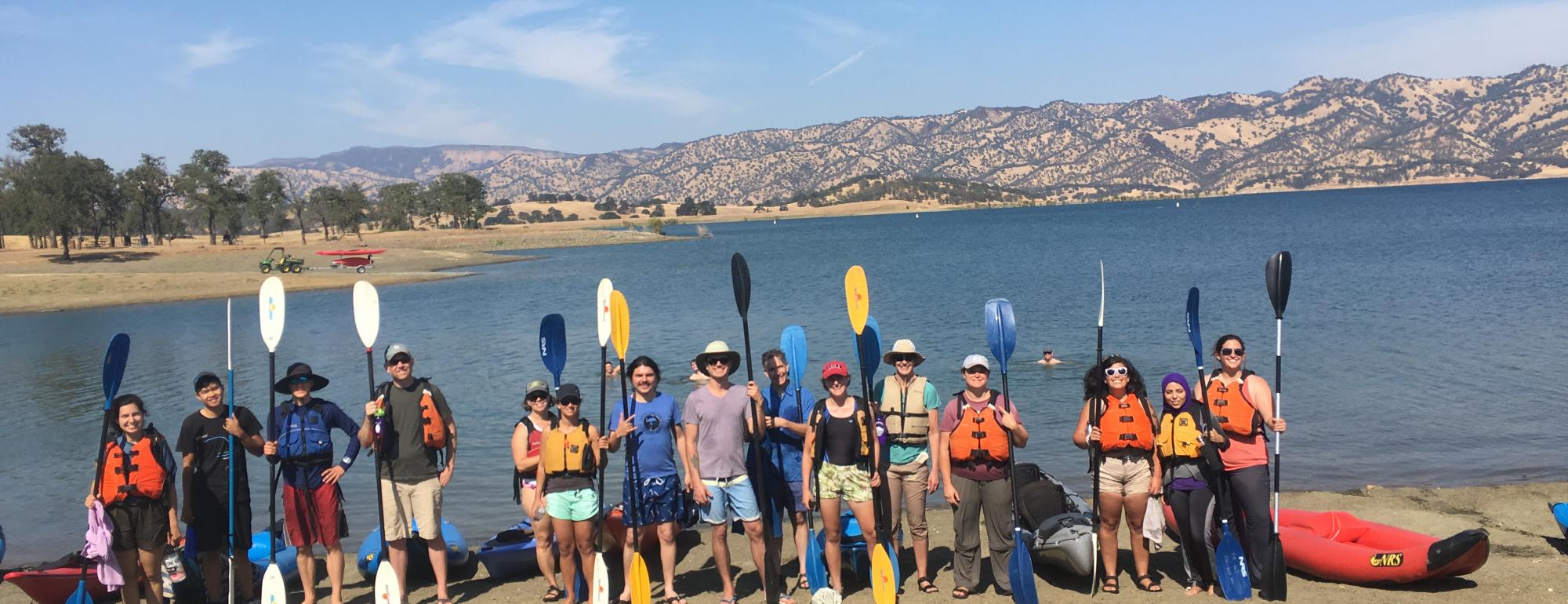 A group of graduate students standing on a lakeshore holding oars and standing next to canoes and kayaks.