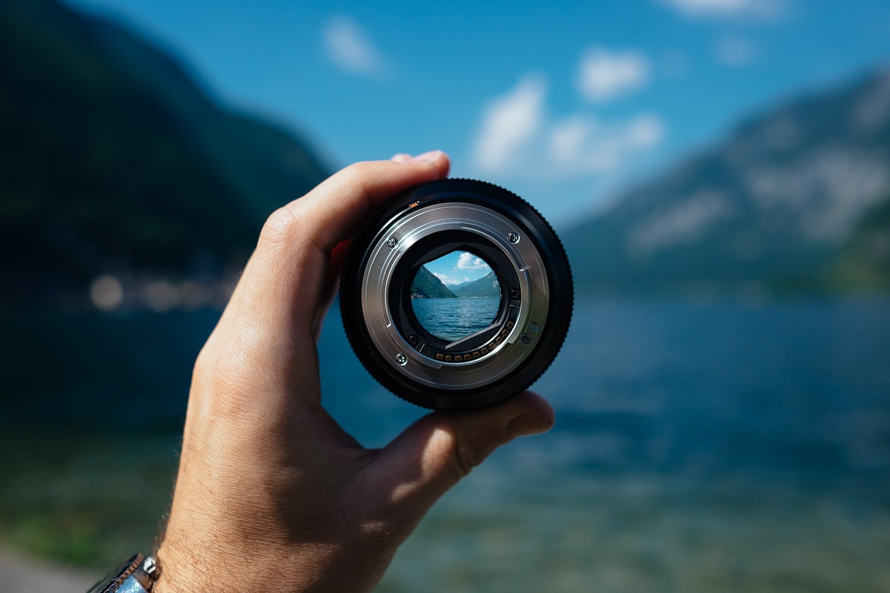 A person's hand hold up a camera lens through which you can see a crisp view of the ocean and steep hillsides while the area outside the lens is blurred.