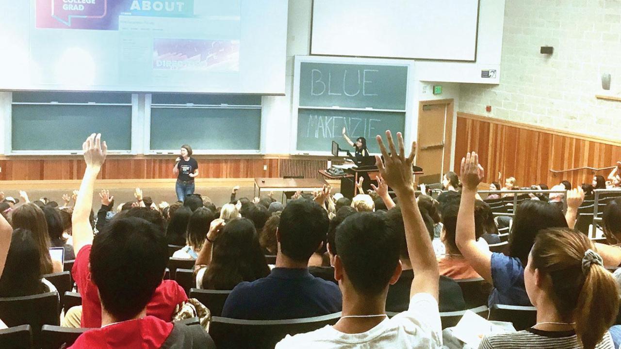 An auditorium with students raising their hands while looking down the room toward the teacher who stand before a wall-sized blackboard.