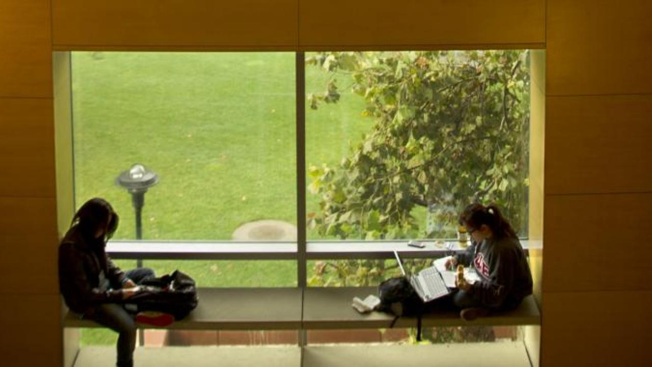 Two students sitting in a window seat, a large bay window behind them overlooking a tree and lawn.