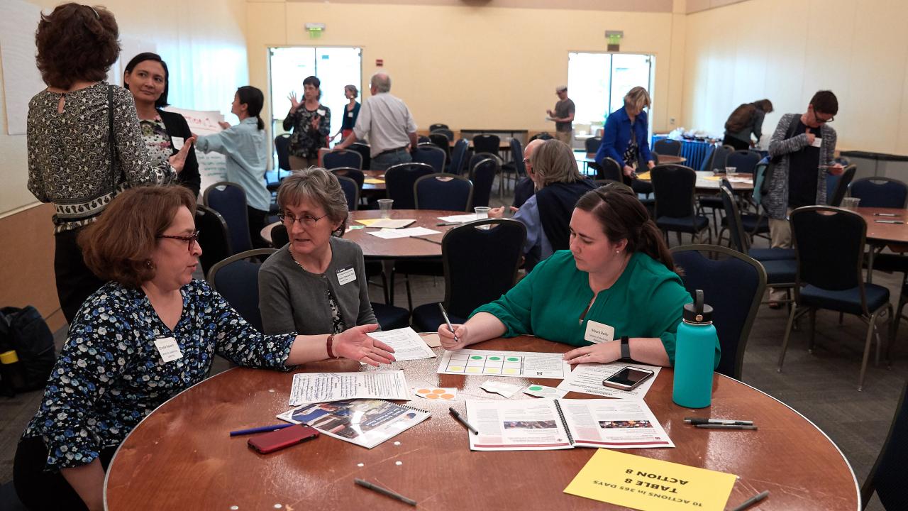 A group of people speaking together at a round table in a conference type venue.