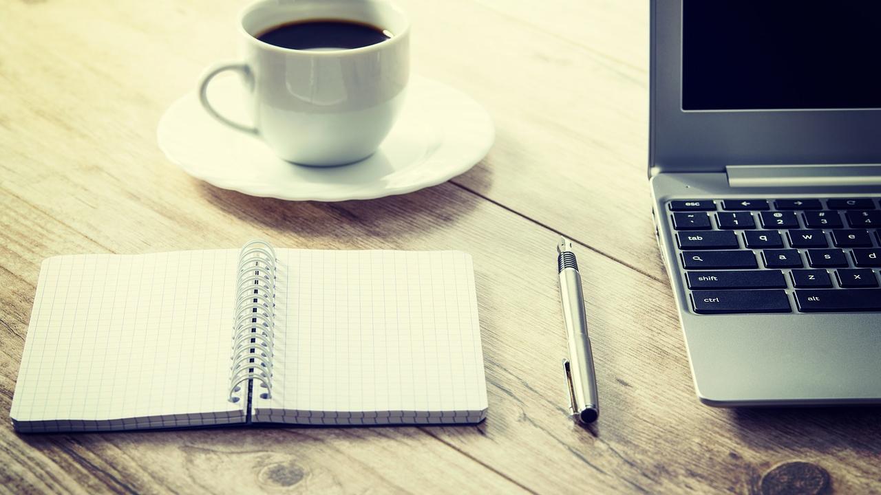 A cup of coffee, an open notebook with a pen, and the side of a laptop are visible on a wooden table.