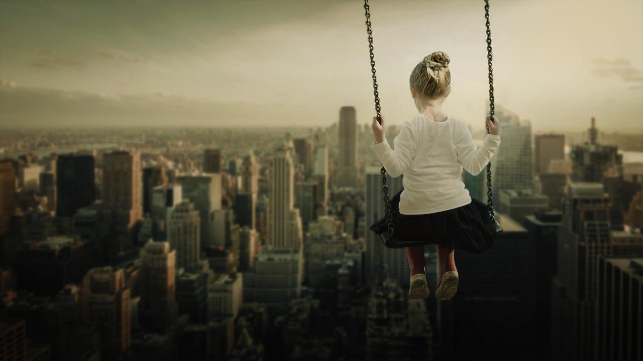 A sepia toned photo of a little girl on a swing with the unusual perspective of swinging over the New York skyline.