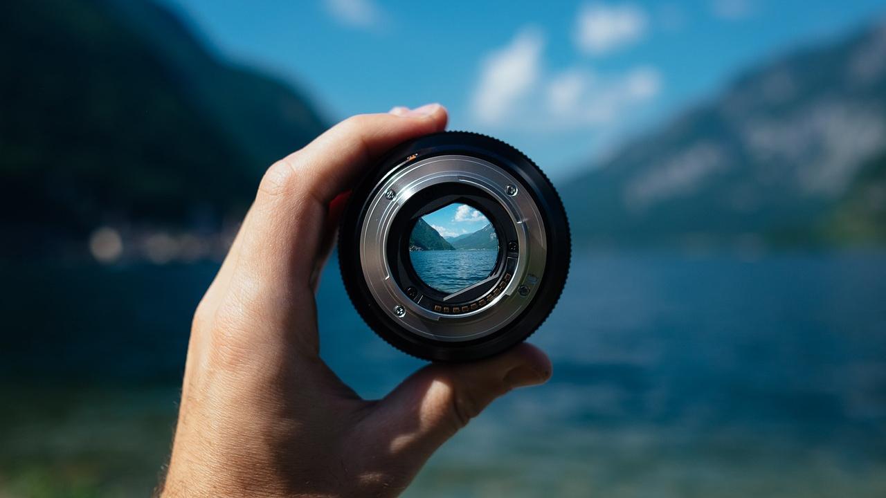 A person's hand hold up a camera lens through which you can see a crisp view of the ocean and steep hillsides while the area outside the lens is blurred.