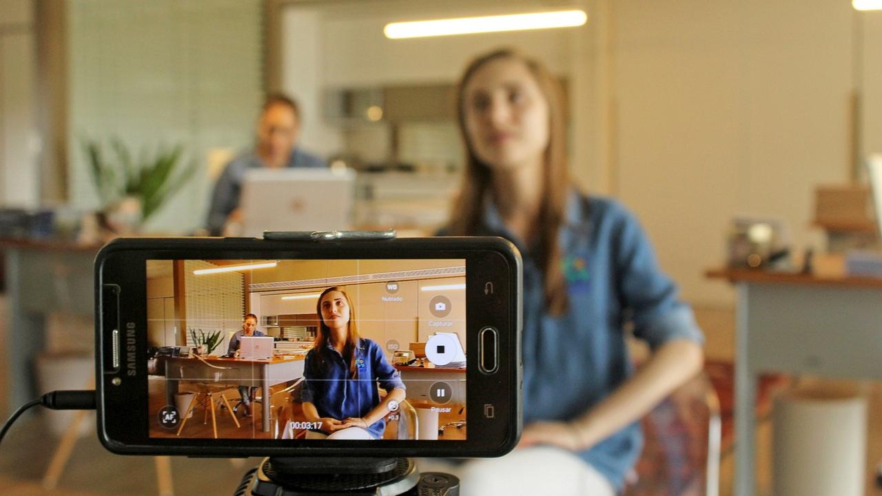A smartphone on a tripod filming a woman at a desk, with another person in the background.