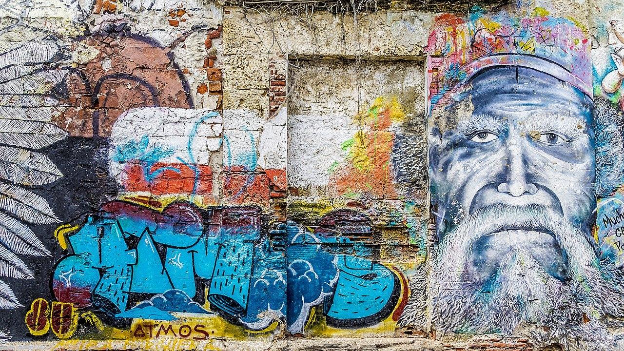 A colorful wall covered in graffiti art, tagged with lettering on the left and center, and the face of a man on the right. Photo by ShonEjai on Pixabay.