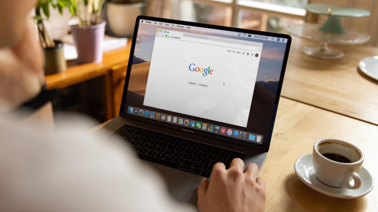 A view over a person's shoulder at a laptop screen whose browser is set to Google search. The laptop sits on a wooden desk with a cup of coffee next to the laptop.