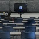 A large classroom where at the front a big screen shows a person speaking from a remote location.
