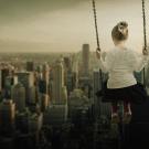 A sepia toned photo of a little girl on a swing with the unusual perspective of swinging over the New York skyline.