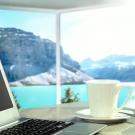 A laptop and coffee sit in the foreground. Beyond is a large window overlooking a lake with mountains in the background.