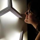 A woman adjusts an illuminated sculpture on the wall in an otherwise dark room, its light softly outlining her face.