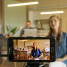 A smartphone on a tripod filming a woman at a desk, with another person in the background.