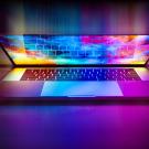 A slightly closed laptop lid exuding a brilliant color display of an abstract pattern and the screen lightly reflecting the keyboard keys.