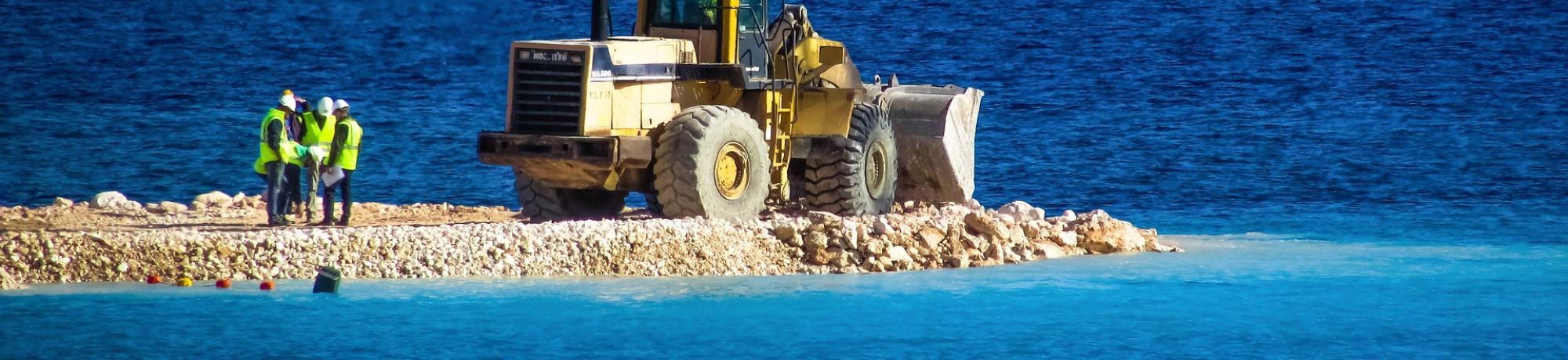 Three construction workers conferring next to a bulldozer, all of which stand on a small man-made gravel island, surrounded by the sea.